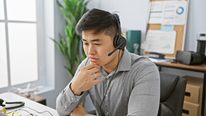 A contemplative young asian man wearing a headset in a modern office setting, embodying professionalism and focus.