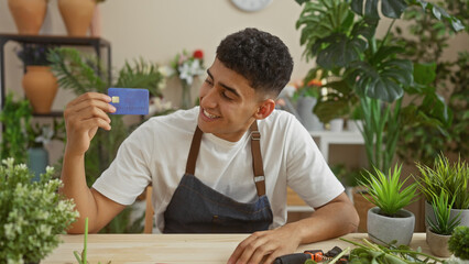 A smiling young man in a flower shop holds a credit card, surrounded by plants and tools.