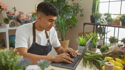 Young man working on laptop at flower shop surrounded by various plants and flowers, creating a natural and vibrant workspace.