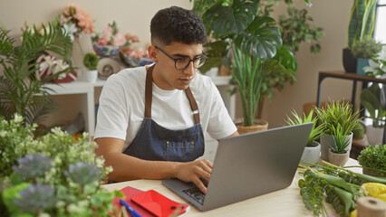 A young man in an apron uses a laptop in a vibrant florist shop surrounded by fresh plants and flowers.