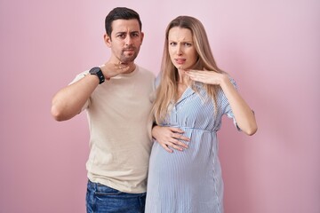 Young couple expecting a baby standing over pink background cutting throat with hand as knife, threaten aggression with furious violence