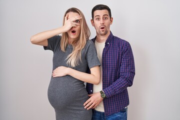 Young couple expecting a baby standing over white background peeking in shock covering face and eyes with hand, looking through fingers with embarrassed expression.