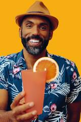 happy bearded mid adult african american man wearing Hawaiian shirt and hat smiling offering orange juice cocktail at viewer on yellow background