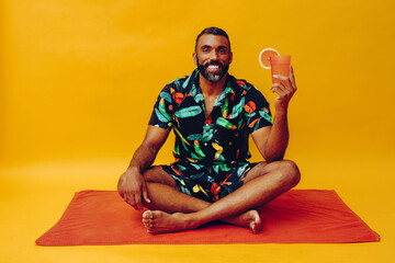 handsome bearded mid adult african american man smiling on vacation sitting on an orange towel, holding orange juice looking at camera