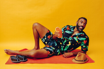 handsome bearded mid adult african american man smiling on vacation lying on an orange towel, holding orange juice looking away