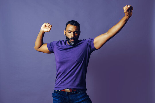 portrait of a successful winner man with a beard and a purple t-shirt celebrating with arm up on a purple background