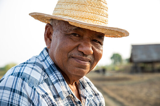 Portrait of elderly farmer man wearing a straw hat standing on dry agricultural area and looking at the camera