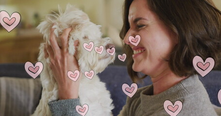 Multiple pink heart icons floating against caucasian woman kissing her dog at home