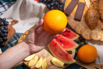 Close-up of hand holding orange against a background of laid out picnic food, selective focus. The concept of summer outdoor recreation on the weekend