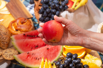 Close-up view of a hand holding a apple against the background of laid out picnic food, selective focus. The concept of summer outdoor recreation on the weekend