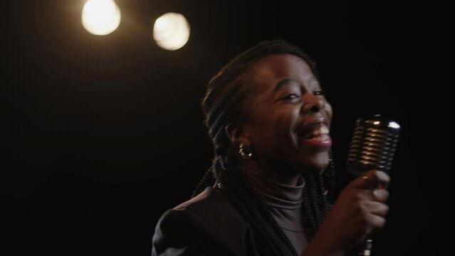 Medium slowmo of joyful young Black female standup comedian going on dark stage and greeting audience in mic at beginning of comedy performance
