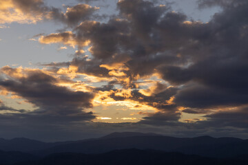 Sunset over a layered mountain range with dramatic dark clouds rimmed in orange light, beautiful...