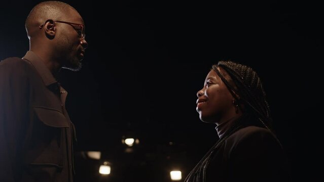 Low angle slowmo of young Black couple looking affectionately at each other while standing on dark stage under projector light during auditions for play at theatre