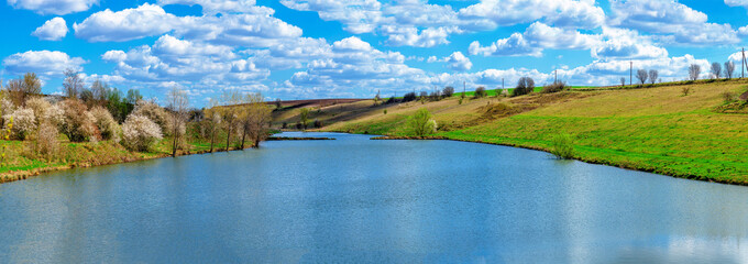 Amazing spring panoramic view with river and green hills, blooming trees. White clouds, blue sky.