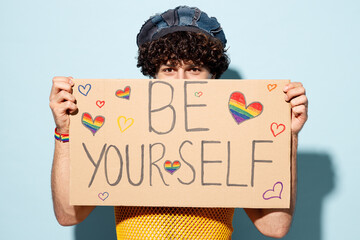 Young cool gay man he wears mesh tank top hat clothes hold cover mouth with card with be your self title text isolated on plain blue background studio portrait. Pride day June month love LGBT concept.