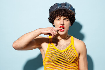 Young happy gay Latin man wear mesh tank top hat clothes with red lipstick make up biting finger isolated on plain pastel light blue background studio portrait. Pride day June month love LGBT concept.