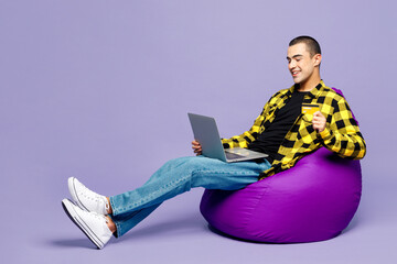 Full body young middle eastern man wear shirt casual clothes sit in bag chair use laptop pc computer hold credit bank card shopping online order delivery book tour isolated on plain purple background.