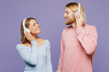 Young fun couple two friends family man woman wear pink blue casual clothes together listen to music in headphones look to each other isolated on pastel plain light purple background studio portrait.