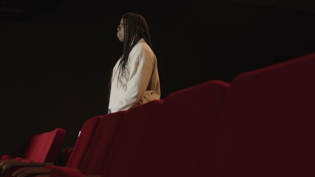 Tracking shot of young Black woman with braided hairstyle and sheets of paper in hands going through empty red theatre seats and climbing on stage to give standup concert