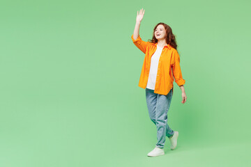 Full body side profile view young smiling happy ginger woman she wear orange shirt white t-shirt casual clothes walk go waving hand isolated on plain pastel light green background. Lifestyle concept.