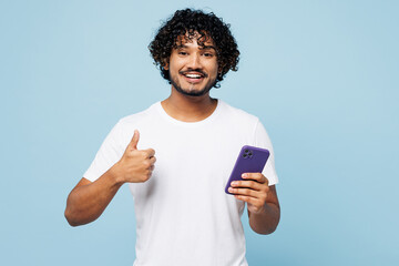 Young happy fun Indian man he wear white t-shirt casual clothes hold in hand use mobile cell phone show thumb up isolated on plain pastel light blue cyan background studio portrait. Lifestyle concept.