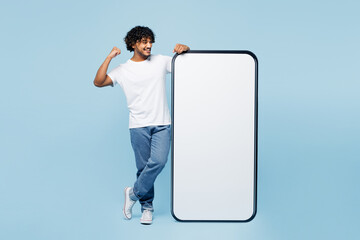 Full body young Indian man wear white t-shirt casual clothes big huge blank screen mobile cell phone smartphone with area do winner gesture isolated on plain pastel blue background. Lifestyle concept.