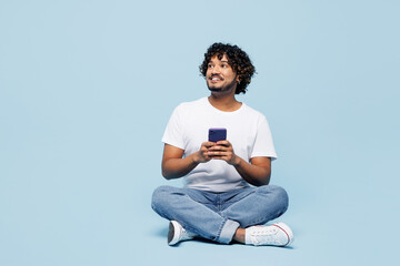 Full body young happy Indian man he wears white t-shirt casual clothes sits hold in hand use mobile cell phone isolated on plain pastel light blue cyan background studio portrait. Lifestyle concept. - obrazy, fototapety, plakaty