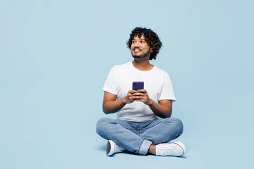 Poster Full body young happy Indian man he wears white t-shirt casual clothes sits hold in hand use mobile cell phone isolated on plain pastel light blue cyan background studio portrait. Lifestyle concept. © ViDi Studio