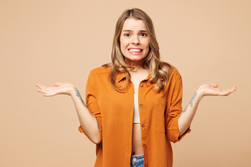 Young confused sad Caucasian woman wear orange shirt casual clothes spread hands shrugging shoulders looking puzzled isolated on plain pastel light beige background studio portrait. Lifestyle concept.