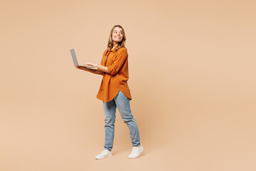 Full body side view young IT woman wear orange shirt casual clothes hold use work on laptop pc computer look aside isolated on plain pastel light beige background studio portrait. Lifestyle concept.