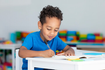 Black primary school boy doing homework, sitting at desk in classroom interior. Training at school
