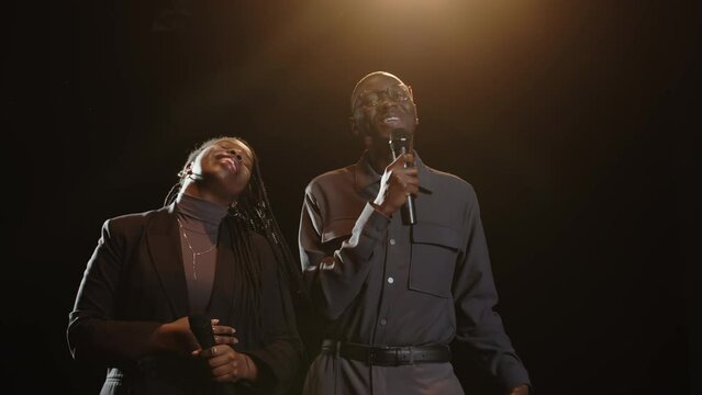 Medium slowmo of young Black couple of standup comedians standing with microphones on dark stage in spotlight and bowing down at camera after performance