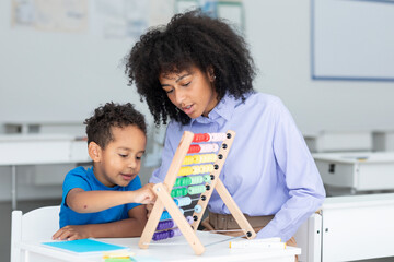 Black female teacher and little boy sitting at desk doing math using abacus, early education concept