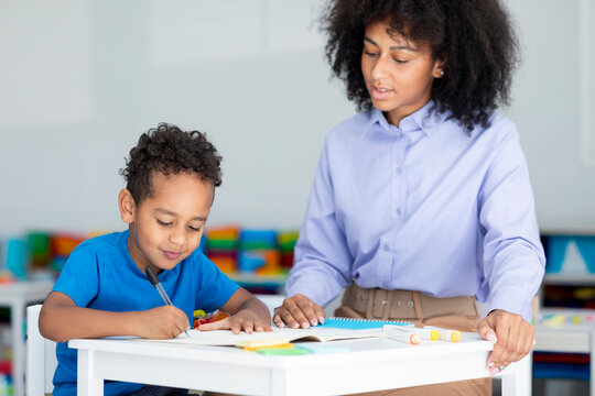 Black teacher woman teaching primary school boy, sitting at desk in classroom, writing and drawing