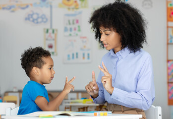 Little black boy learning to count on fingers with female teacher help, sitting at desk in classroom interior