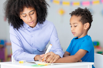 Little smart black boy studying with female tutor and writing in notebook, sitting at desk in...