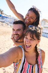 Vertical. Group of cheerful diverse young gen z friends taking a selfie on the beach, smiling and having fun together doing the piggyback. People enjoying summer leisure time and holidays travel