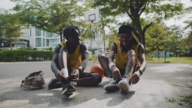 Wide shot of two African American sportsmen tying shoelaces on their sneakers sitting on streetball playground before game