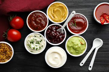 Different tasty sauces in bowls, tomatoes and spoons on black wooden table, flat lay