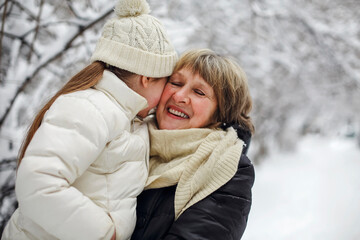 Loving grandmother with little girl granddaughter on snowy weather outdoors, smiling senior woman lovingly straightens knitted hat on little girls head while walking in winter park. Selective focus