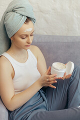 Young woman hold creme jar on couch, head wrapped in towel on head
