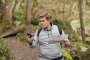 A young man tries to find a phone signal in the forest. Searching for mobile connection in wild nature on vacation. Angry man tourist hiker holding smart phone and paper map - obrazy, fototapety, plakaty