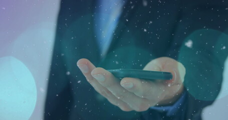 Image of snow falling and light spots over caucasian businessman using smartphone