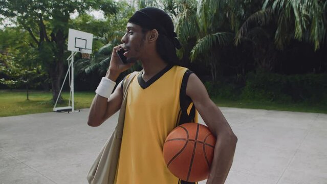 Handheld shot of male streetball player calling his friend on mobile phone on outdoor playground
