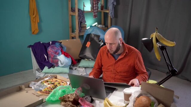 Millennial man anxious desperate worker annoyed work crumpling sheet of paper throw away working in messy home living room with scattered stuff. Mess, disorder and interior concept.