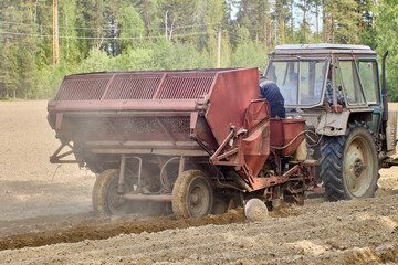 Potato planter is hitched to an agricultural wheeled tractor during spring sowing in plowed farmer field.