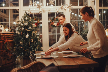 Mother, father and daughter setting festive Christmas table together at cozy decorated rustic...