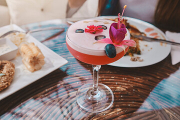 A glass of pink drink with a flower on top sits on a table next to a plate, Food and drink