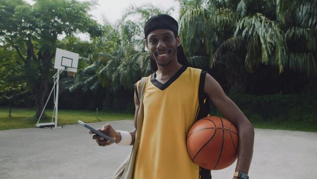 Medium portrait of male African American athlete holding basketball ball, checking smartphone and smiling at camera on court
