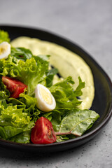 Fresh green salad with quail eggs, cherry tomatoes and guacamole in black plate on grey background